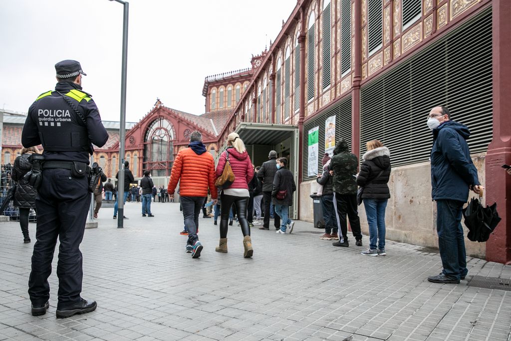 Votants que fan cua a l'exterior del Mercat de Sant Antoni i que esperen el seu torn per entrar a votar, i un agent de la Guàrdia Urbana que fa tasques de vigilància durant el dispositiu