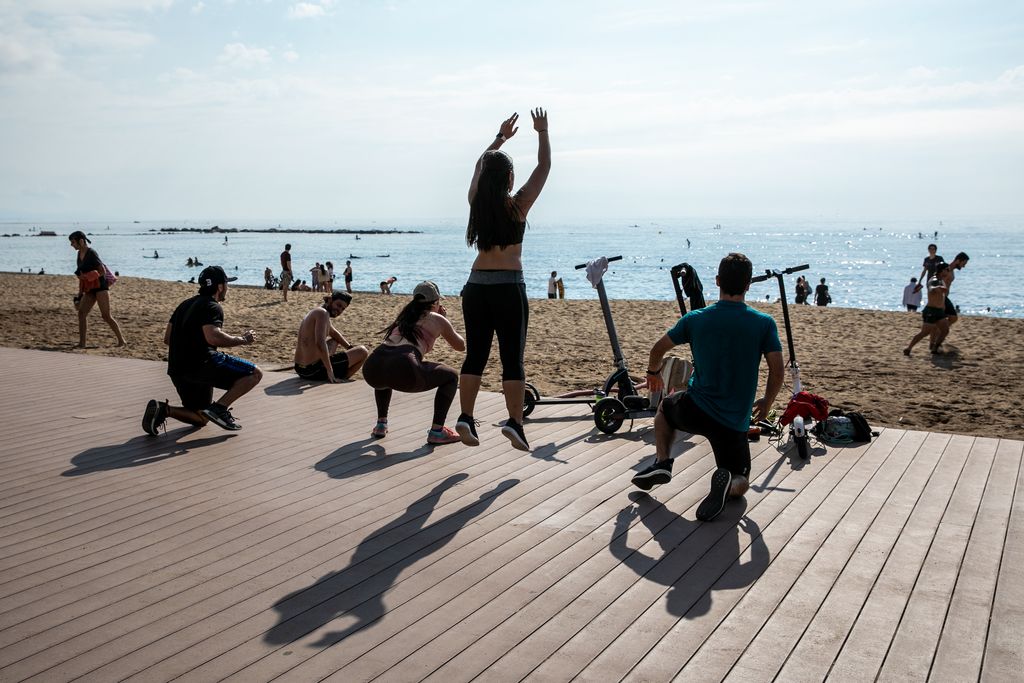 Grup de persones fent exercicis gimnàstics a la platja i, al fons, banyistes i gent practicant surf de rem