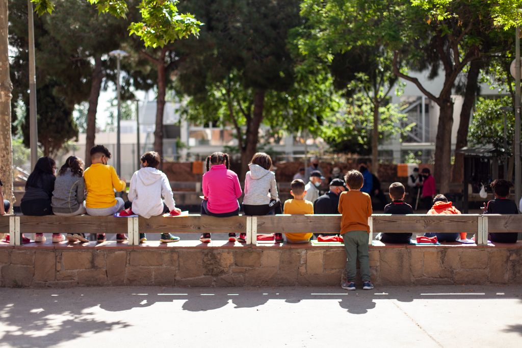 Nens de l'Escola Mercè Rodoreda asseguts en uns esglaons de la plaça de Nou Barris amb els apunts a les mans assistint a una classe