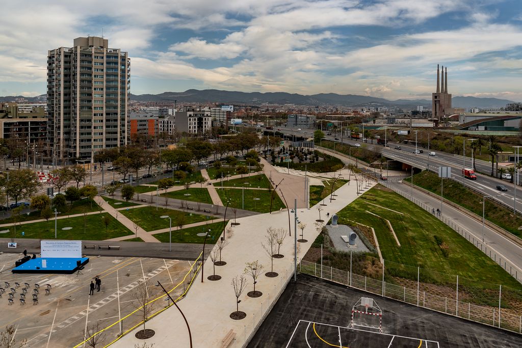 Fotografia elevada de la zona del parc Campus Besòs amb un passeig central i una pista per a bàsquet i futbol, un passeig central i zones verdes amb l'àrea de joc infantil al fons. Darrere, es veu el final de la ronda del Litoral i la Central Tèrmica de Sant Adrià