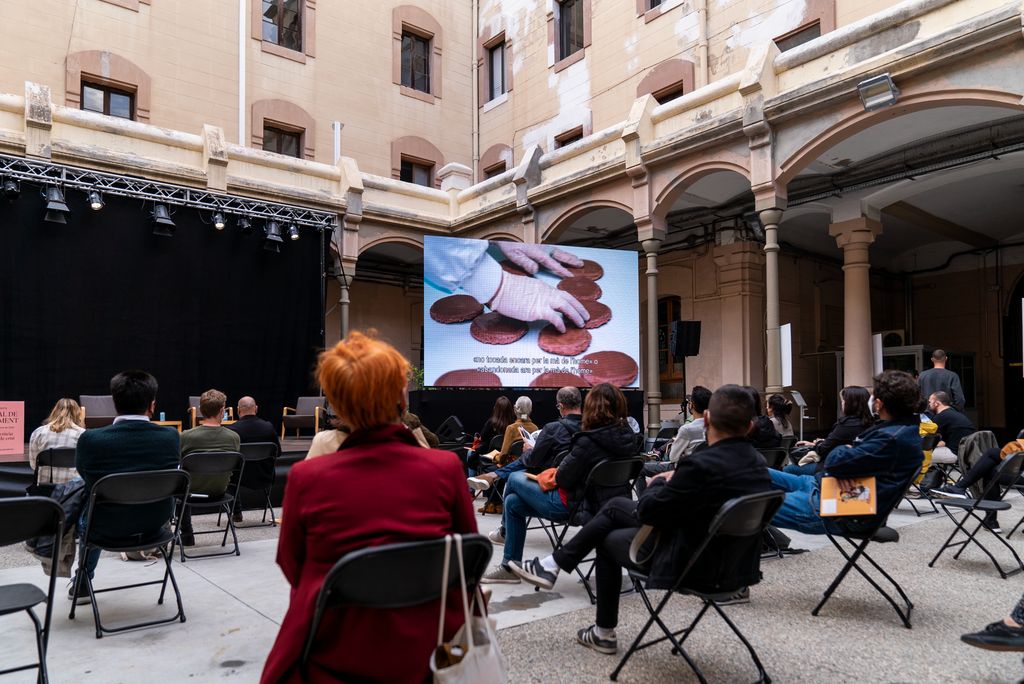 Vista de l'escenari i d'una gran pantalla de la Biennal de pensament a la Model abans de l'inici de la taula rodona "La ciutat del futur" amb el públic assistent ocupant els seus seients