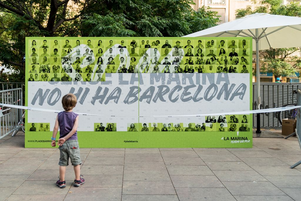Pla general del mural fotogràfic “SENSE LA MARINA NO HI HA BARCELONA” amb un nen mirant-s’ho durant la presentació del Pla de barris