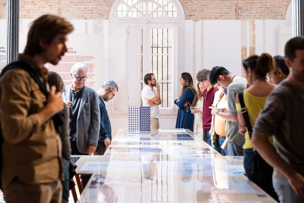 Visitantes de la exposición “99+ Imaginarios / Las Barcelonas futuras” contemplando las obras expuestas en unas grandes mesas protegidas con cristal en la Fundación Enric Miralles