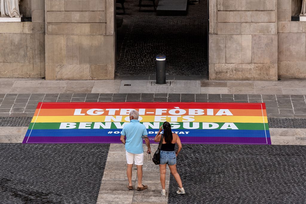 Pancarta contra la LGTBIfobia en el suelo ante la entrada del Ayuntamiento de Barcelona, con personas observándola en la plaza de Sant Jaume