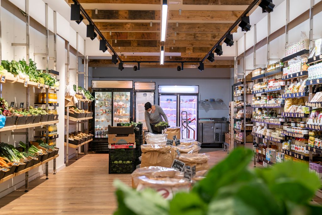 Botiga de queviures ecològics on la venedora mou les caixes de carxofes acabades d’arribar. A un costat es veuen prestatges amb fruites i verdures i, a l’altre, aliments empaquetats. Al centre hi ha un refrigerador per als productes que necessiten fred i, als passadissos, uns sacs per a la venda al detall de fruita seca i llegums.
