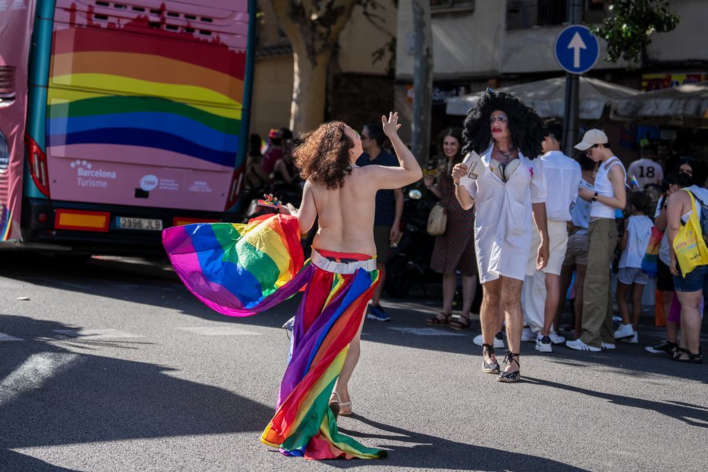 Una persona sense samarreta, i amb una faldilla i una bandera amb els colors LGTBI, durant la manifestació del Pride! 2023. De fons, una persona vestida d’infermera.