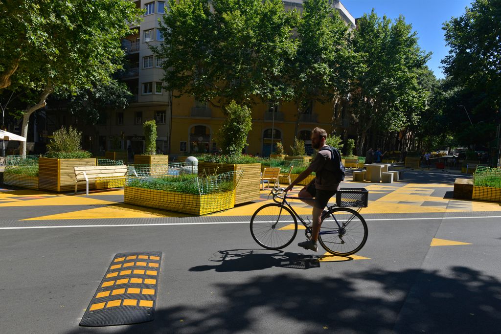 Un ciclista circula per una zona pacificada de la superilla de Sant Antoni amb una zona delimitada per al pas dels vehicles dels veïns amb ressalts per evitar l’excés de velocitat. Darrere seu es veu una zona delimitada per jardineres i senyalització horitzontal groga destinada al lleure