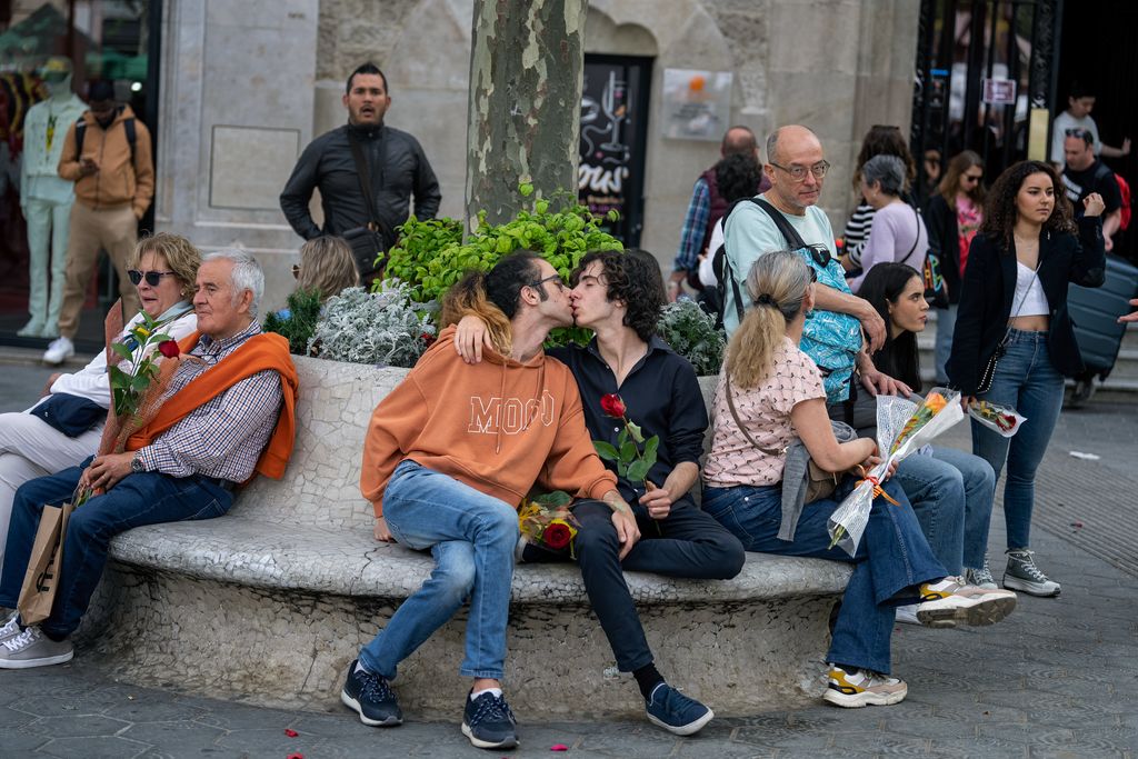Una pareja, sentada en un banco del paseo de Gràcia, se da un beso durante el Día de Sant Jordi 2023. A su alrededor, varias personas pasean.