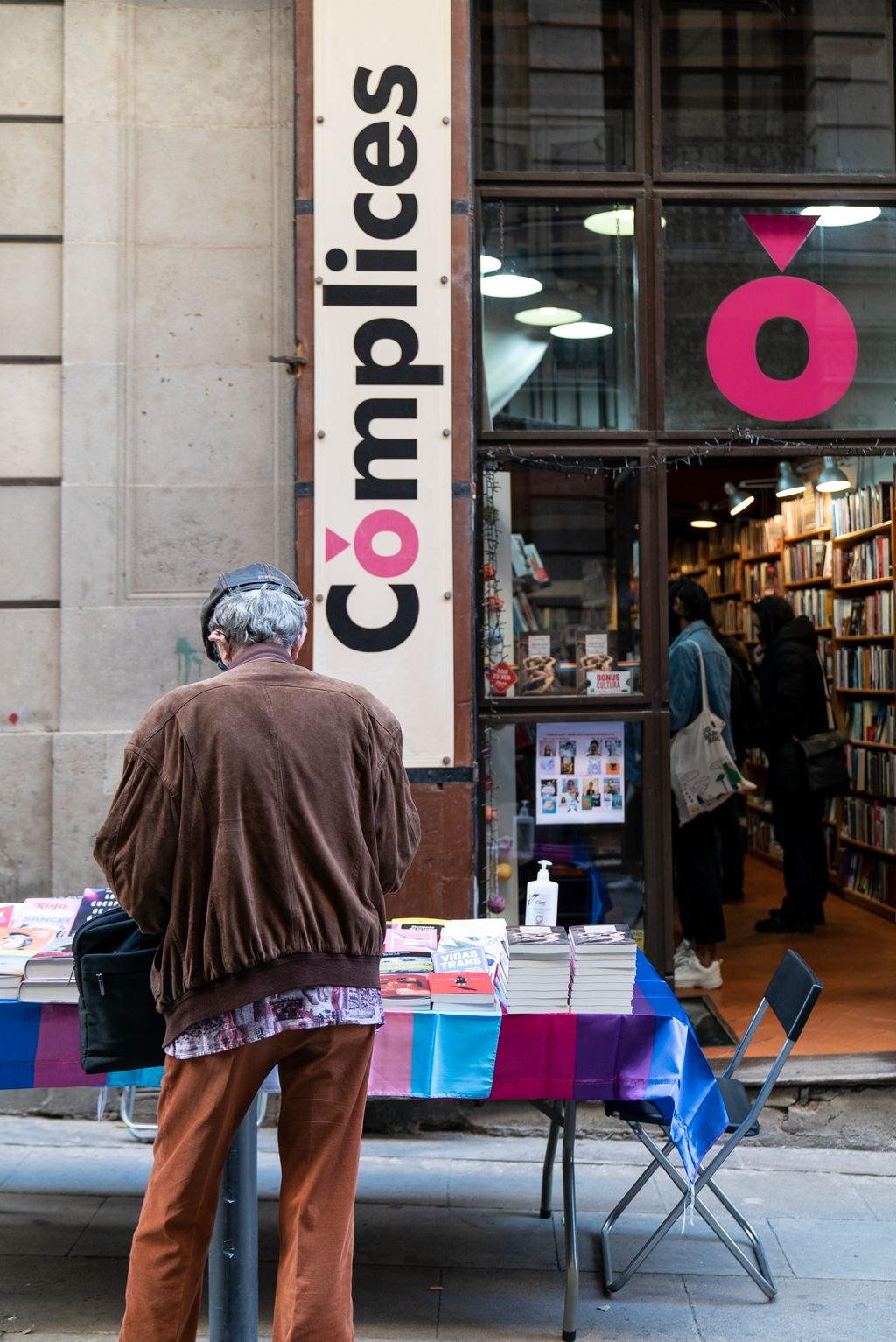 Un hombre mirando los libros del puesto delante de la librería Cómplices montado con motivo del Día de Sant Jordi. En el interior de la tienda también se ven clientes comprando y hojeando libros