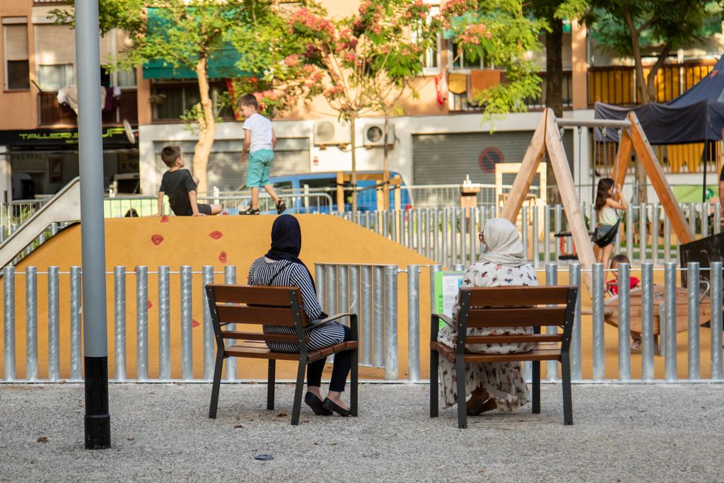 Unas madres con hiyabs sentadas en los bancos observan a sus hijos jugando en la zona de juego infantil de los jardines de la Mediterrània