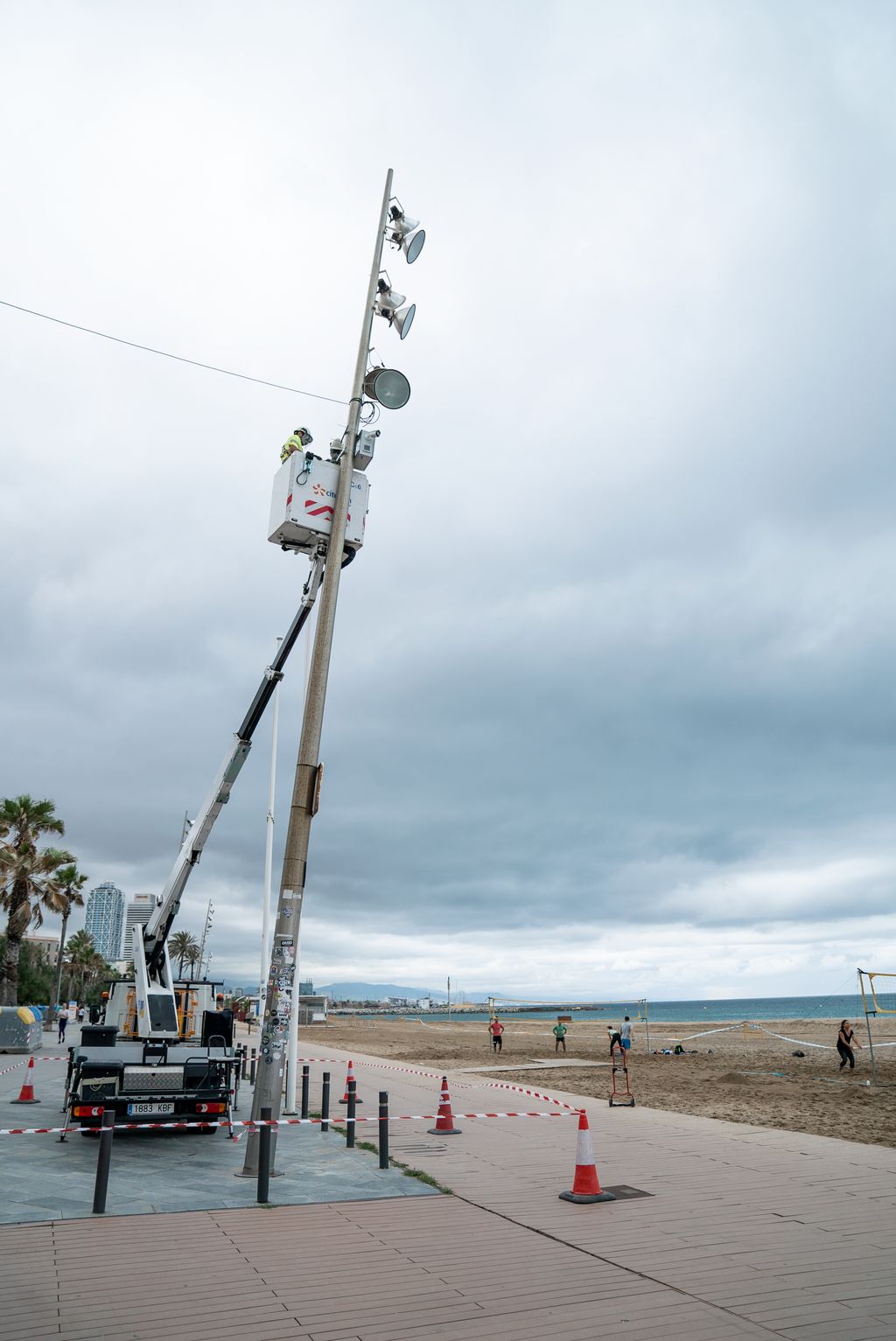Un treballador enfilat en una grua amb elevador posa les càmeres amb sensors per controlar la capacitat dalt d'un fanal a la platja. Al voltant, la zona està precintada com a zona de treball