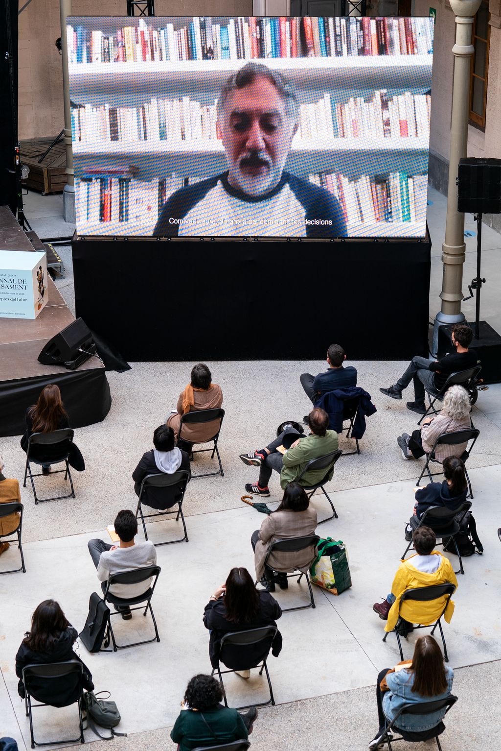 Vista zenital sobre el públic assistent a la taula rodona "La ciutat del futur" a la Model i de la pantalla on apareix un dels convidats, l'arquitecte Hashim Sarkis