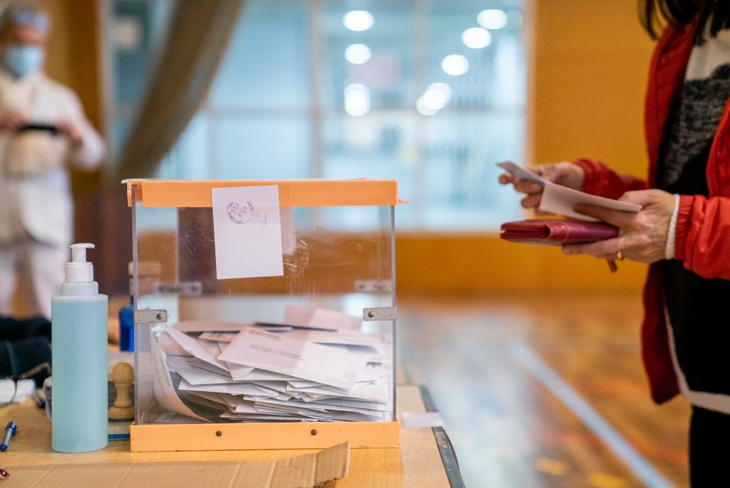 Una dona es troba davant d'una de les urnes del CEM Guinardó i prepara el vot i la documentació. L'urna està segellada i precintada per evitar qualsevol manipulació. Al fons, hi ha una persona vestida amb un equip de protecció individual