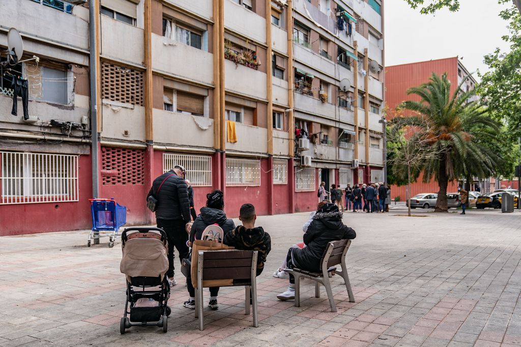 Veïns del Besòs asseguts en uns bancs es miren un grup de persones aturades davant de la porta d’un edifici