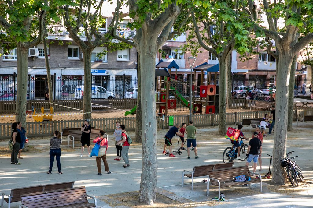 Vista del parc de l'Espanya Industrial, a prop del carrer de Muntadas, on hi ha grups d'adults conversant i de nens jugant. L'àrea de jocs infantils roman tancada
