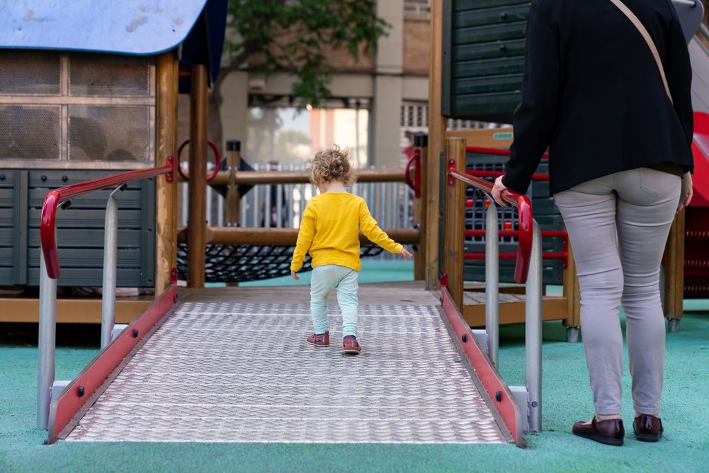 Una niña pequeña sube por la rampa de un espacio de juego infantil en los Jardines de Elx bajo la mirada de su madre