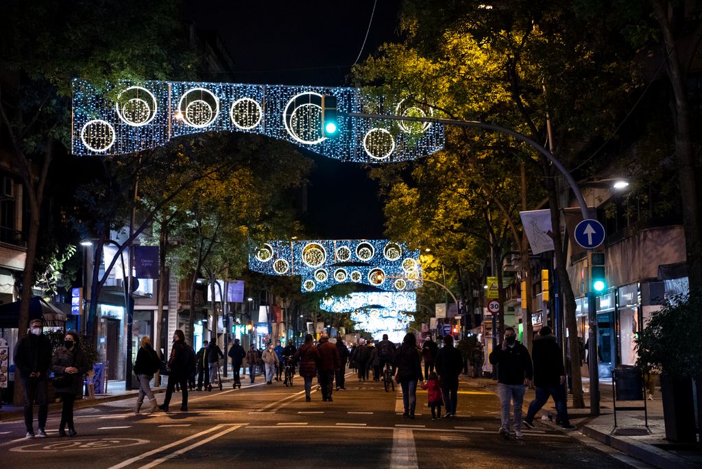 Gent passejant pel carrer de la Creu Coberta tancat al trànsit durant l’“Obrim carrers” i gaudint de la il·luminació de Nadal amb mosaics de llums blaves i esferes de color blanc i groc