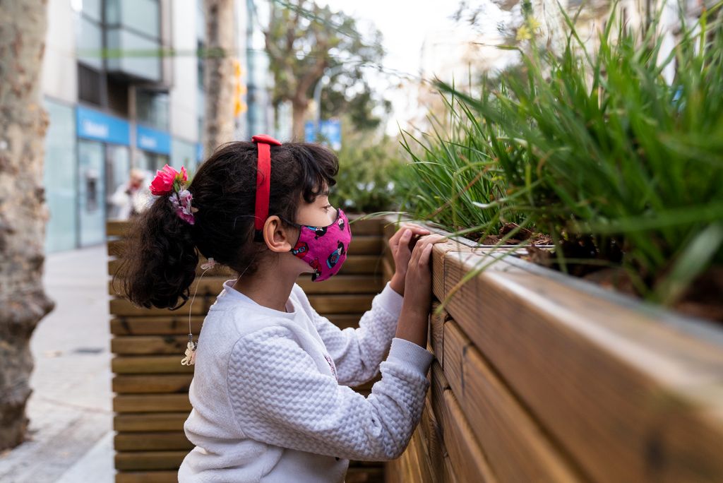 Una nena amb mascareta mira les plantes de les jardineres instal·lades per separar del trànsit la zona de vorera ampliada davant de la porta de l’Escola Carlit
