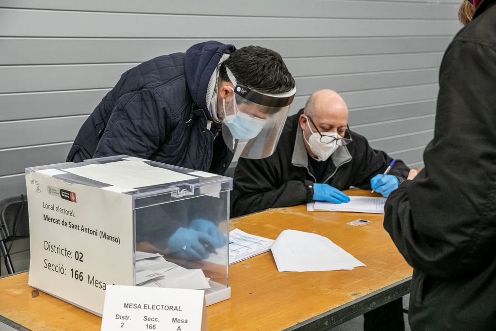 Un membre de la mesa que porta una pantalla protectora revisa un nom al llistat de votants