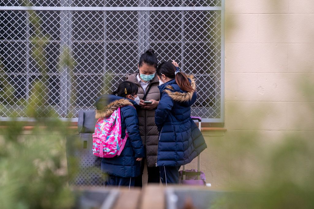 Unas niñas con su madre a la salida del colegio, esperando en la zona pacificada de acera ampliada delante del Colegio Sagrat Cor Ribes. Las tres miran un móvil, y en primer plano están las jardineras altas que separan el área del tráfico