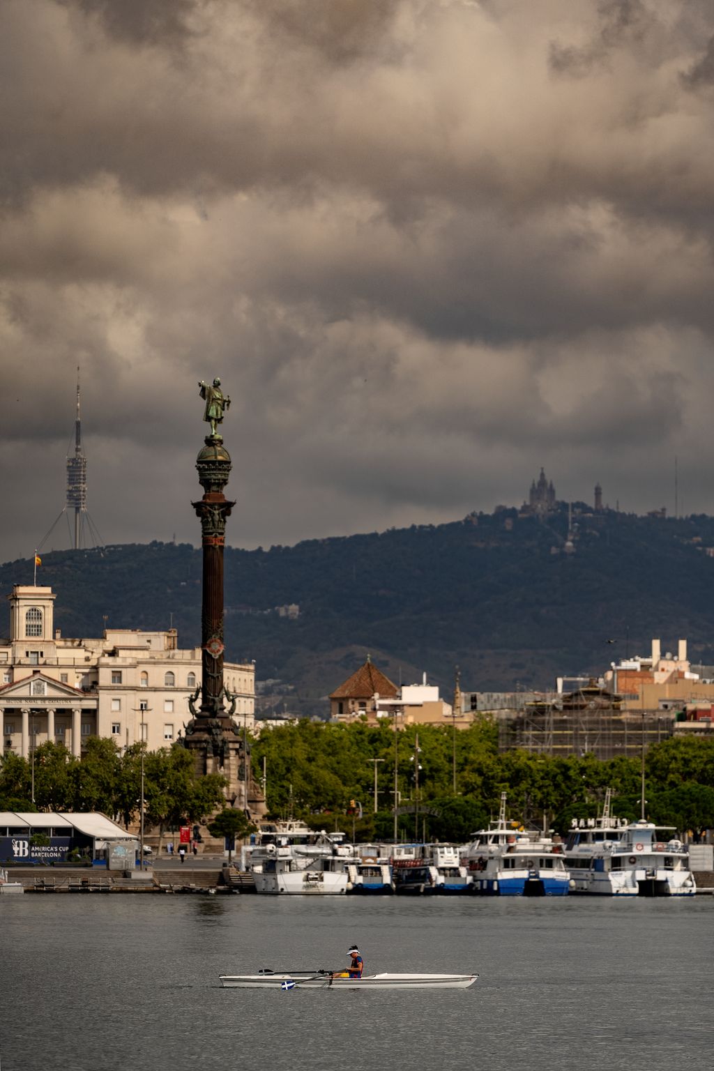 Una persona en una piragua al port. Al fons, l'estàtua de Colom, la Torre de Collserola i el Tibidabo