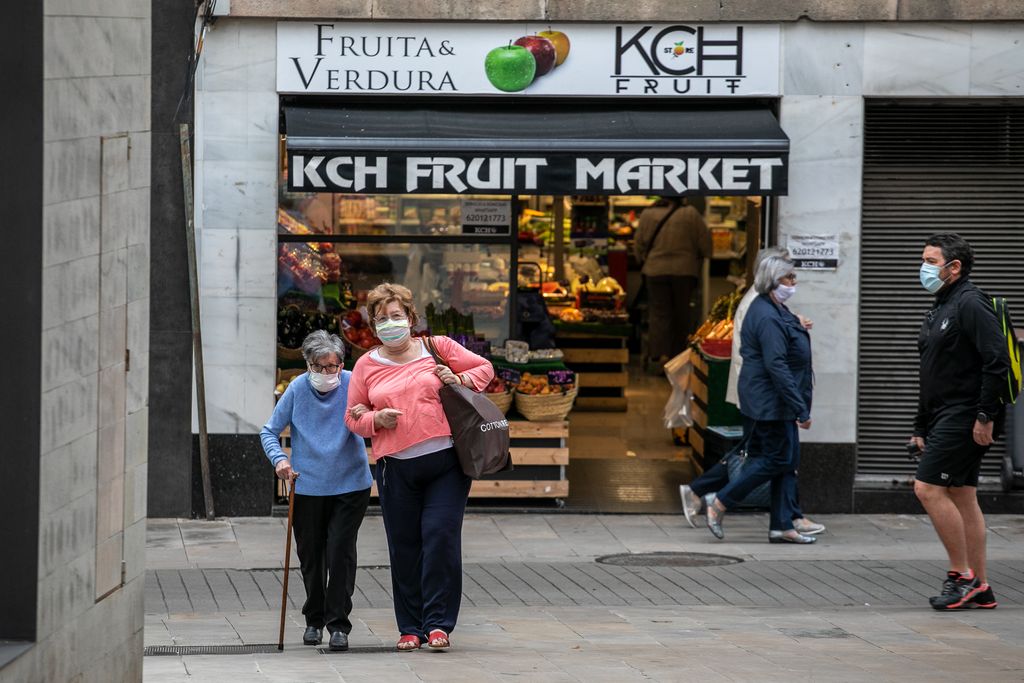 Una dona gran acompanyada d'una altra caminen amb una bossa de la compra prop d'una fruiteria al carrer Major de Sarrià