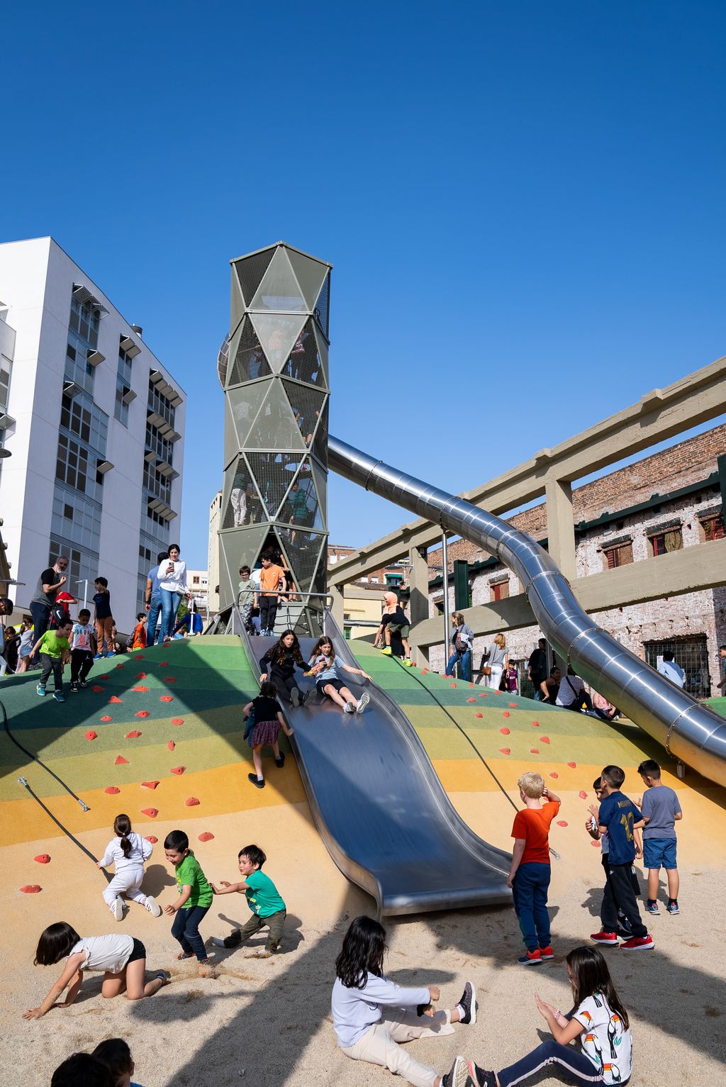Niños y niñas y personas adultas en la zona del tobogán del área de juegos de Can Batlló.