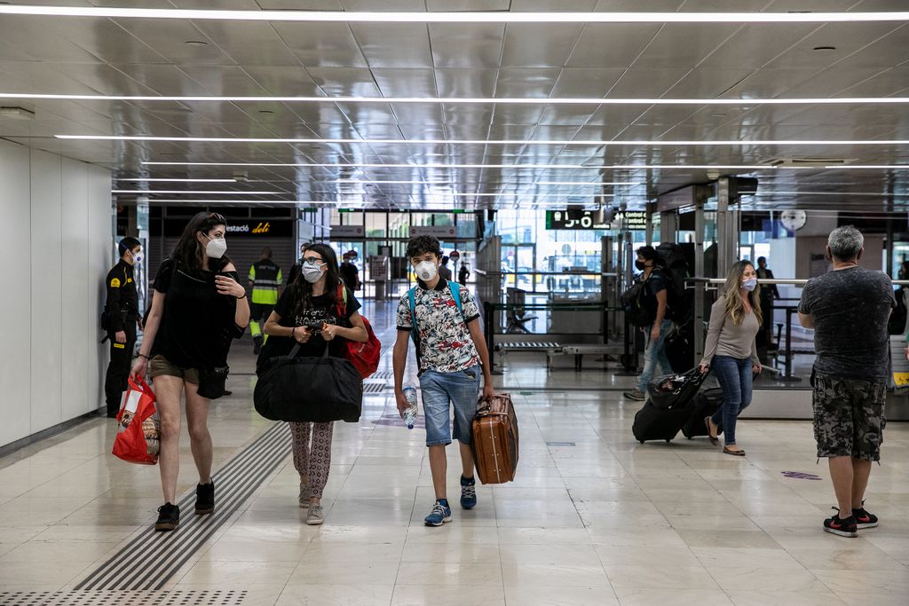 Gent dins de l'estació de Sants amb maletes i bosses de viatge, amb les mascaretes posades i passant per davant dels restaurants i botigues tancats