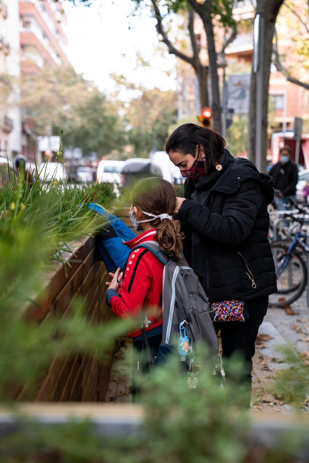 Una nena amb la seva mare al costat mira les plantes de les jardineres instal·lades per separar del trànsit la zona de vorera ampliada davant de la porta de l’Escola Carlit