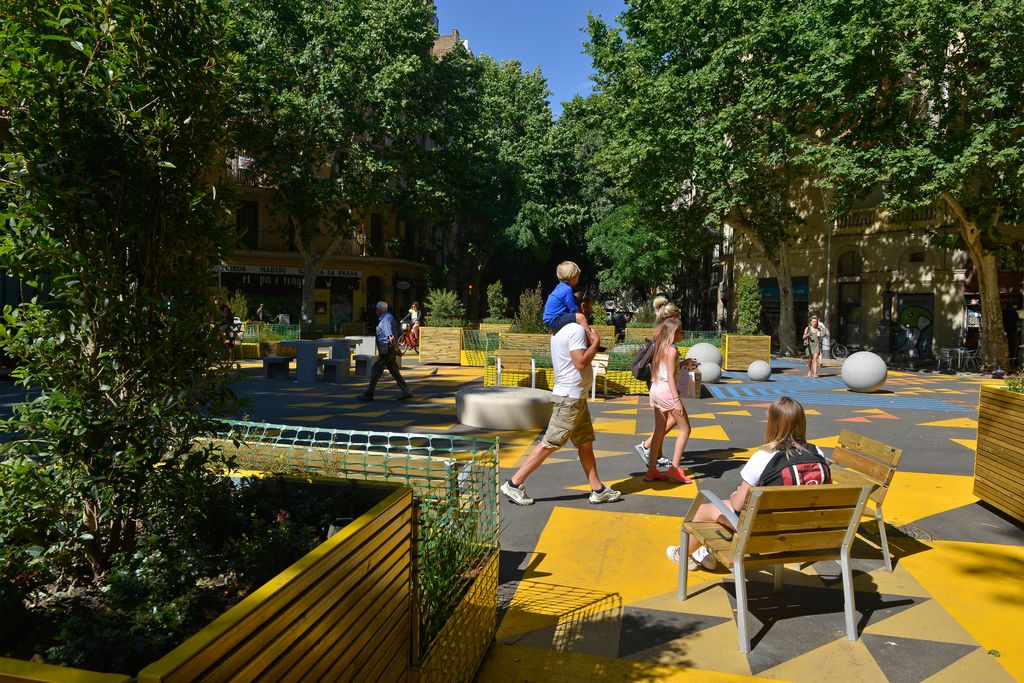 Zona de la supermanzana de Sant Antoni delimitada con jardineras y señalización horizontal amarilla por donde pasa una familia con un niño en brazos. Al lado, una joven sentada en uno de los bancos o asientos individuales