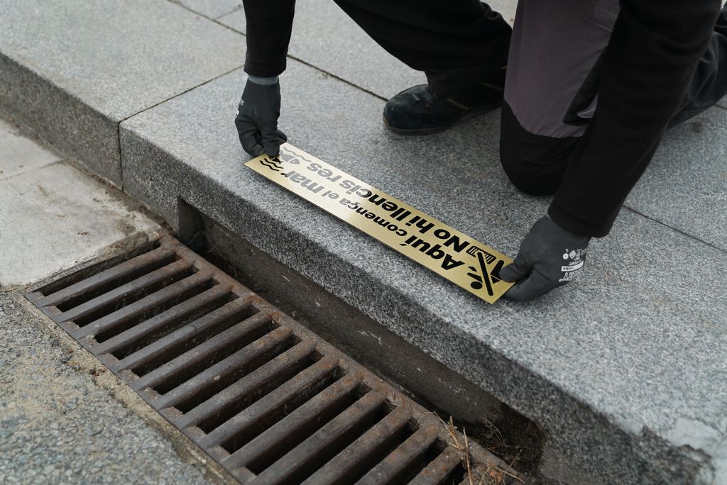 Un treballador col·loca una placa en una vorera del Passeig Marítim, sobre un embornal. A la placa hi diu: "Aquí comença el mar. No hi llencis res"