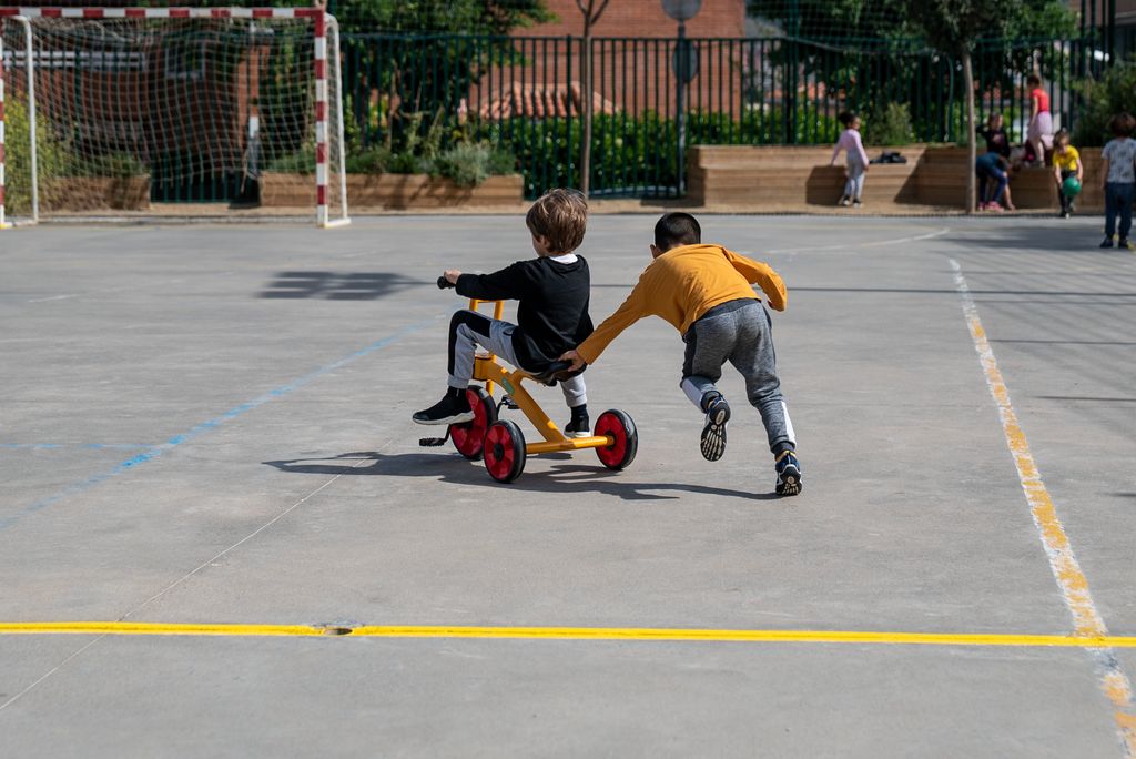 Un nen va en tricicle per la pista de ciment del pati d'una escola mentre un altre l'ajuda