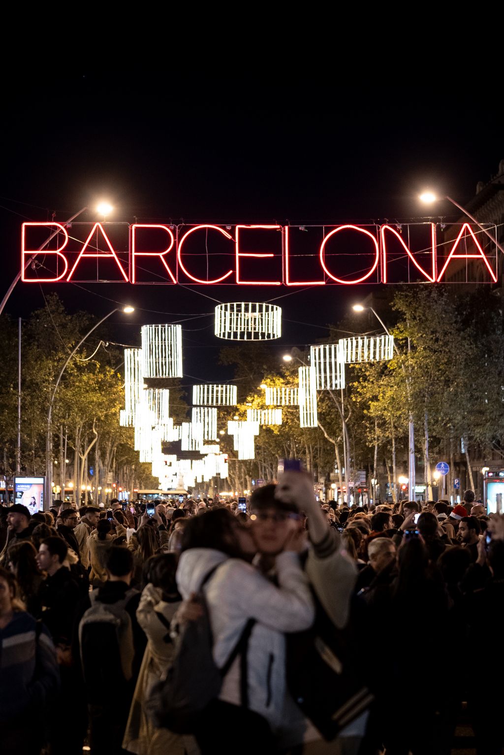 Una pareja haciéndose una fotografía con el móvil. De fondo, multitud bajo las luces de Navidad de Gran Via de les Corts Catalanes con las letras de Barcelona