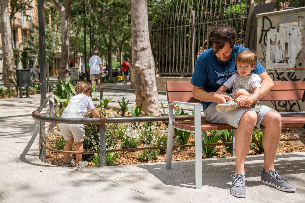 Un home amb un infant a la falda assegut en un banc. Al darrere, un infant
toca la vegetació de l’eix verd de Consell de Cent.