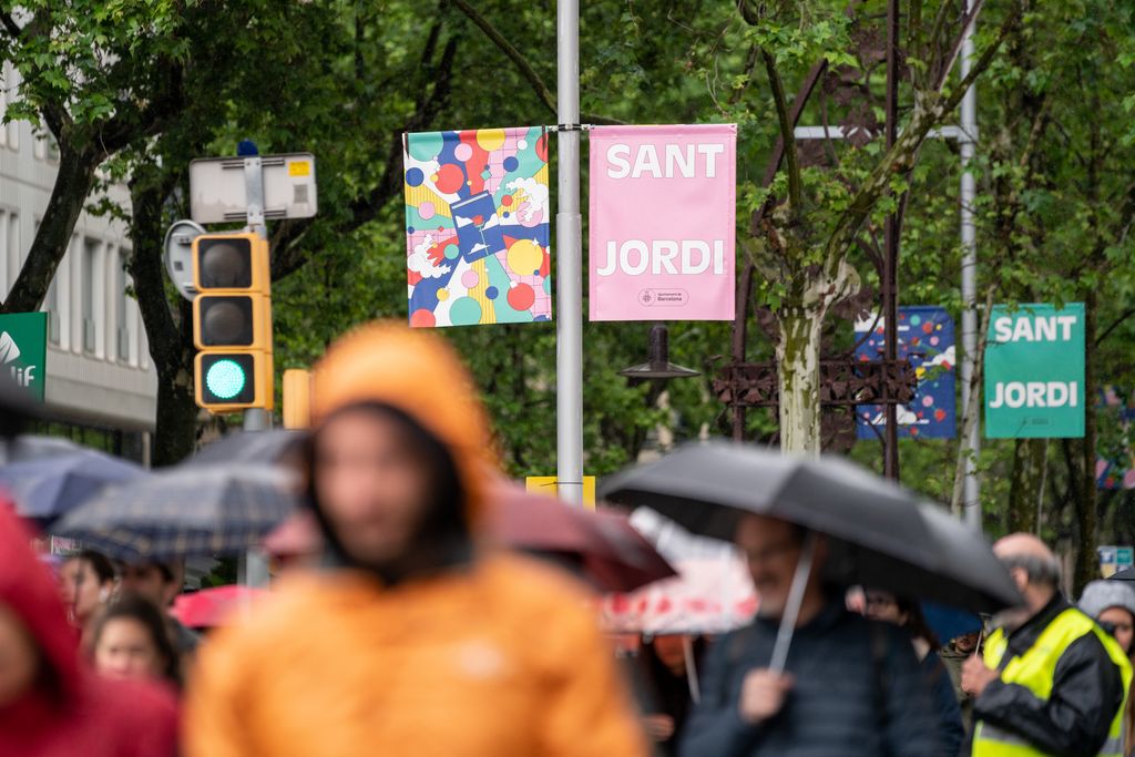 Gent passejant sota la pluja per la superilla literària de Sant Jordi i banderoles de Sant Jordi penjant dels fanals