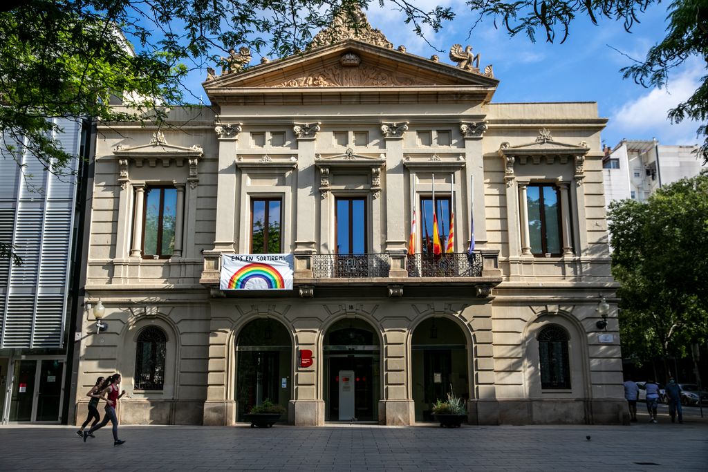 Plaça de Comas amb la pancarta "Ens en sortirem" penjada de la façana de la seu del districte. Dues noies passen corrent per davant