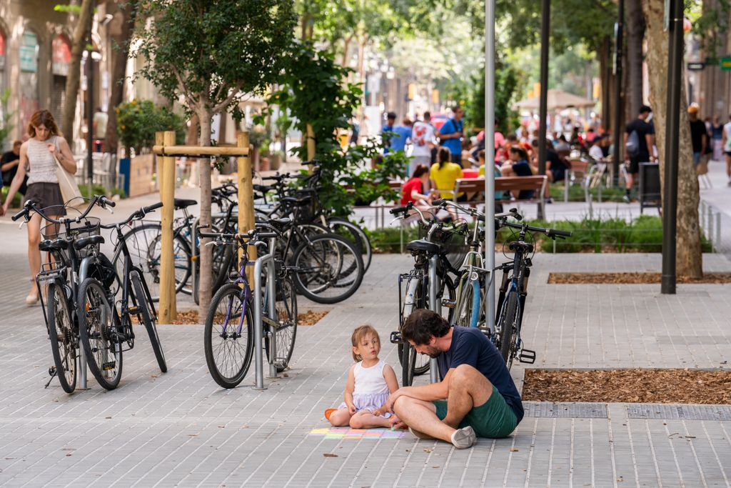 Una nena i un home asseguts al terra de Consell de Cent pintant amb guixos de colors els panots del carrer Consell de Cent. Al darrere, un conjunt de bicicletes aparcades.