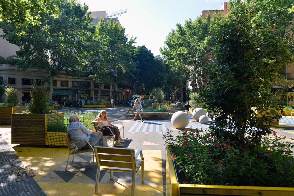 Zona de jocs de la superilla de Sant Antoni delimitada per jardineres i amb el paviment pintat en diferents colors. Hi ha veïns asseguts als seients i nens jugant