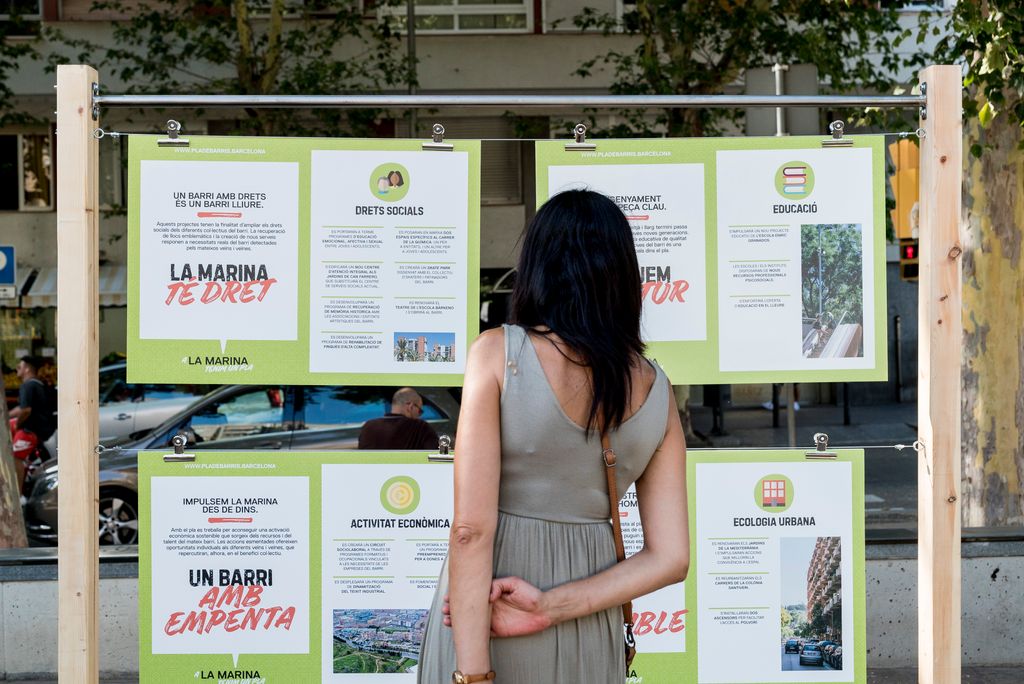 Una dona d’esquena mira cartells penjats en una estructura de fusta on podem llegir, entre d’altres, “LA MARINA TÉ DRET”; “UN BARRI AMB EMPENTA” a la presentació del Pla de barris