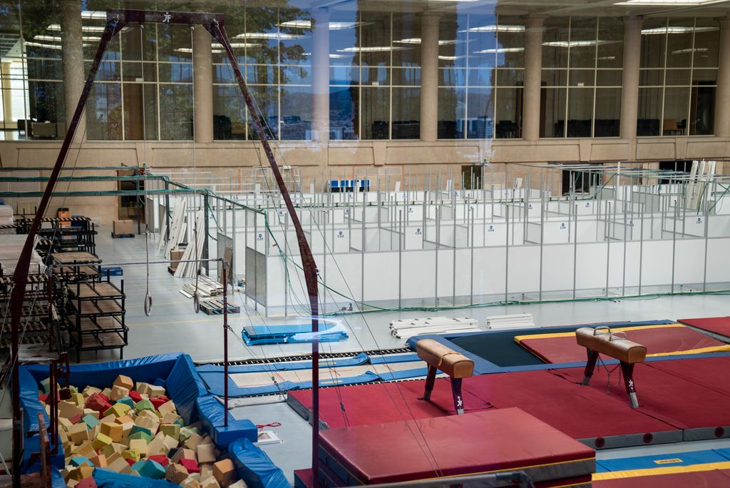 Vista d'una gran sala on l'emplafonat i el material del Pavelló Salut de l'INEFC comparteix espai amb les anelles, la fossa de recepció, trampolins, cavalls i cavalls amb arcs