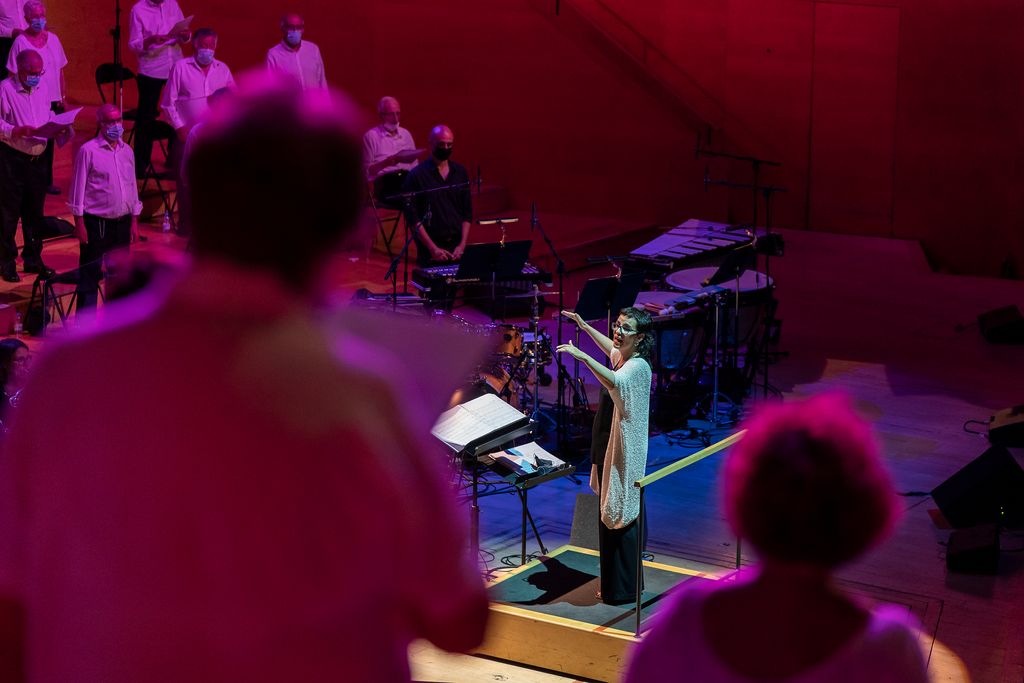 Membres del cor drets cantant a L’Auditori durant la celebració del XX Concert de les Persones Grans de Barcelona “Canta Gran!”, amb la directora al fons
