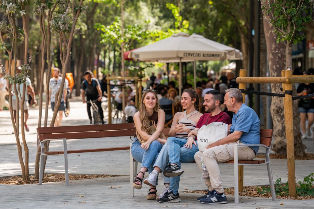 Quatre persones conversen i riuen assegudes a un banc de l’eix verd de Consell de Cent. Al fons, gent passejant.