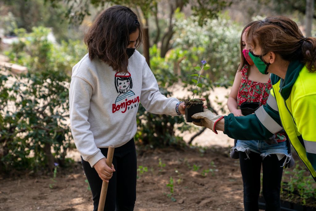 Una jardinera li passa un test amb una flor a una de les nenes participants a la plantada de vegetació al parc del Guinardó perquè la pugui plantar al terra