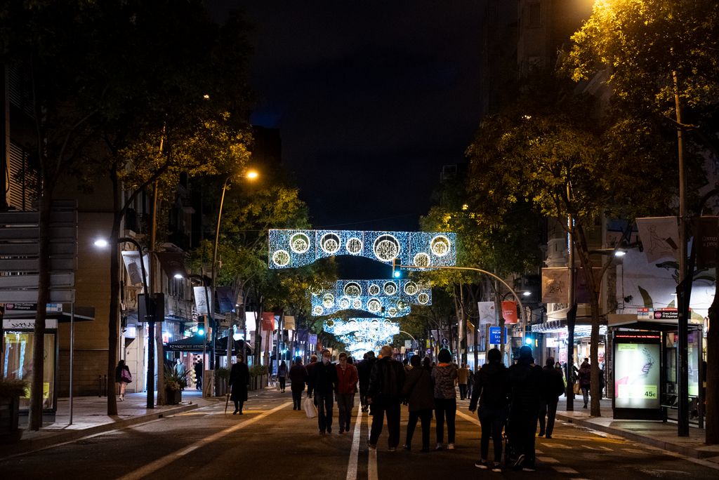 Gent passejant pel carrer de la Creu Coberta tancat al trànsit durant l’“Obrim carrers” i gaudint de la il·luminació de Nadal amb mosaics de llums blaves i esferes de color blanc i groc